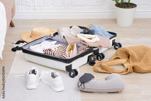 Open suitcase full of usual female clothes, shoes and summer accessories on floor in living room. photo