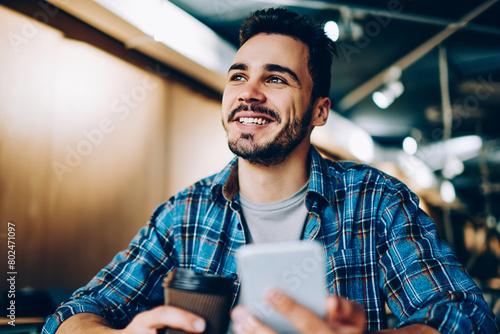 Cheerful handsome guy looking up imagine his vacation while chatting via smartphone in office, young smiling student dreaming about something on coffee break holding mobile phone for networking #802471097