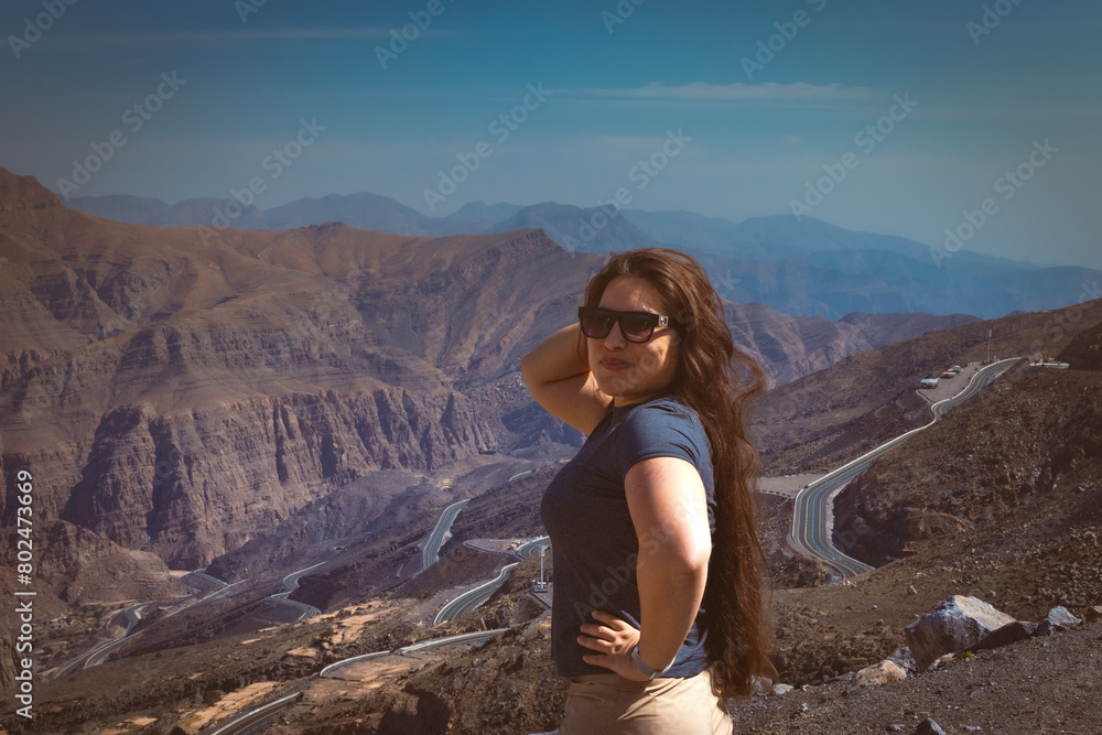 Fototapeta premium Portrait of a happy woman hiker standing on the slope of mountain ridge against mountains, blue cloudy sky on background. The woman is happy and looking at camera. Travel and active lifestyle concept.