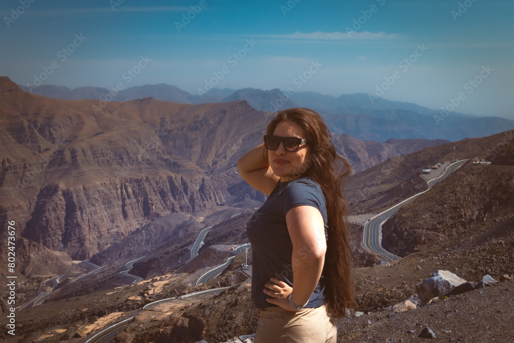 Naklejka premium Portrait of a happy woman hiker standing on the slope of mountain ridge against mountains, blue cloudy sky on background. The woman is happy and looking at camera. Travel and active lifestyle concept.