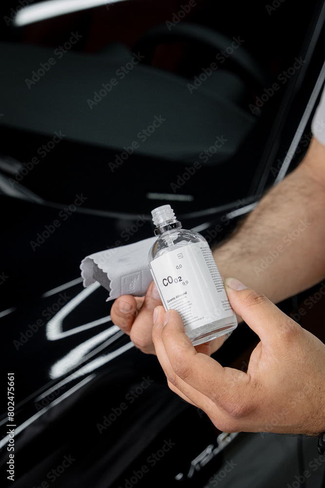 Specialistic ceramic coating liquid poured onto sponge by unrecognizable professional in protective gloves. Car detailing process. Black car in the background.