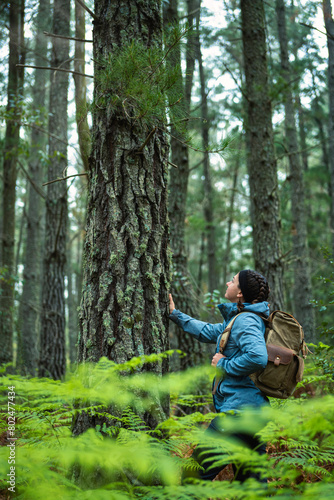 Young woman with backpack touching and observing the trunk of a pine tree, in the foreground ferns out of focus, love for nature and sustainability.
