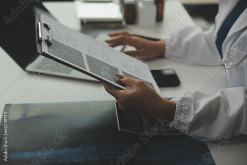 Serious female doctor using laptop and writing notes in medical journal sitting at desk. Young woman professional medic physician wearing white coat and stethoscope working on computer at workplace.