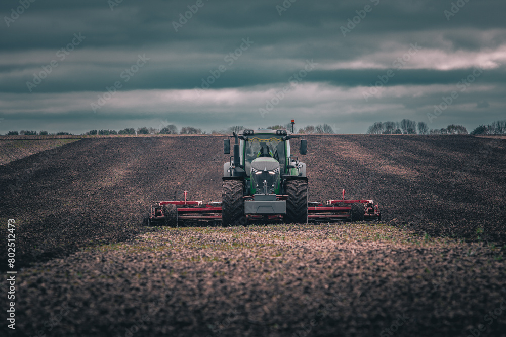 Fototapeta premium Tractor with cultivator working on a field under dark moody sky