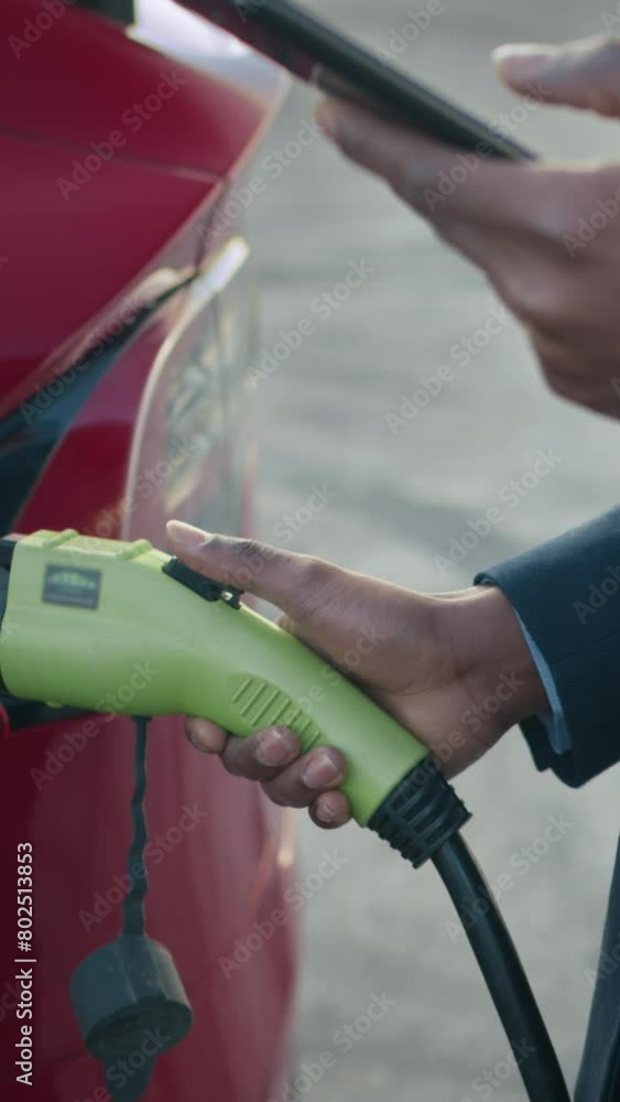 Vertical Screen: African american man in suit using smartphone while standing outdoors with cable from charge for electric car. Concept of modern technology. Man using smart phone connect to electric Vertical Screen: African american man in suit using smartphone while standing outdoors with cable from charge for electric car. Concept of modern technology. Man using smart phone connect to electric