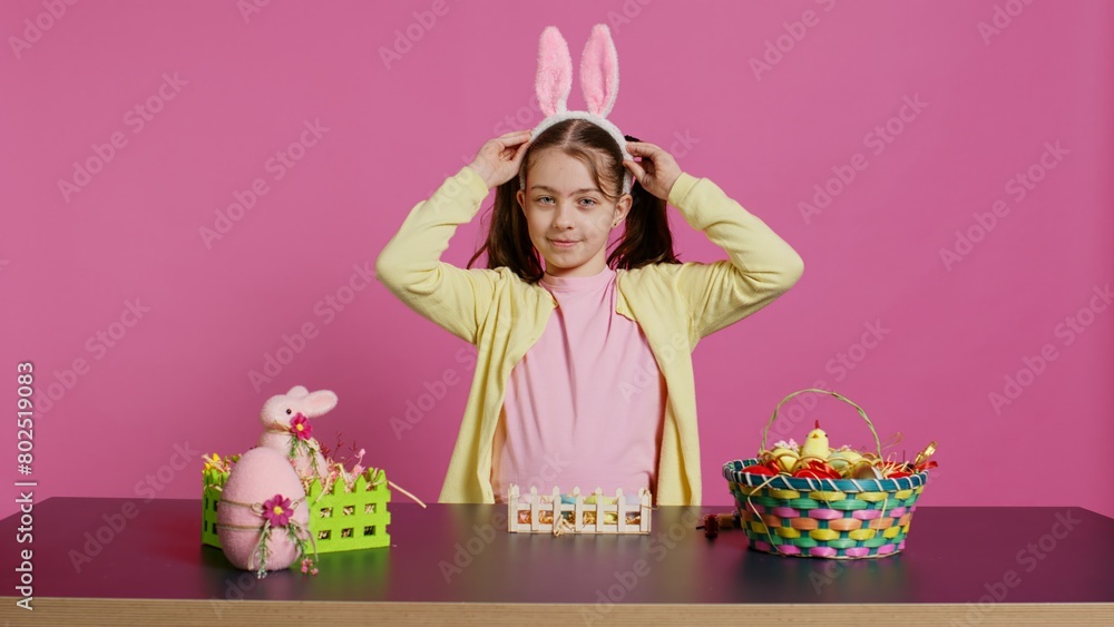 Energetic young girl with adorable bunny ears waving in studio, saying hello and greeting someone while she creates easter decorations. Joyful toddler posing against pink backdrop. Camera B. Energetic young girl with adorable bunny ears waving in studio, saying hello and greeting someone while she creates easter decorations. Joyful toddler posing against pink backdrop. Camera B.