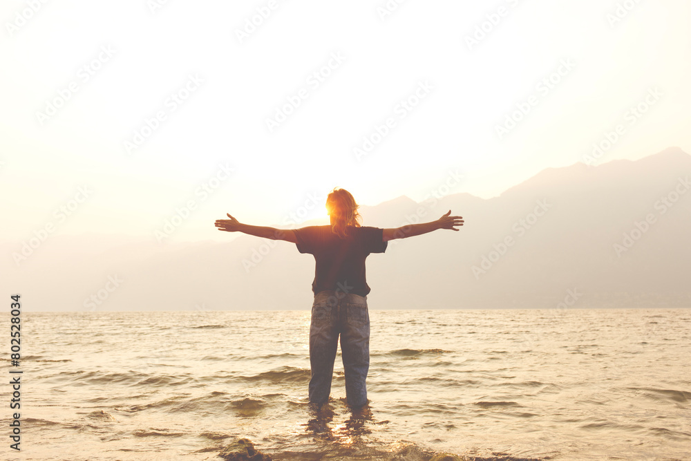 Fototapeta premium woman with open arms welcomes the warmth of the light of the setting sun on the banks of a lake