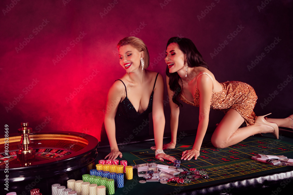 Photo of two charming laughing ladies wear sexy dresses enjoying playing roulette isolated red fog background Photo of two charming laughing ladies wear sexy dresses enjoying playing roulette isolated red fog background