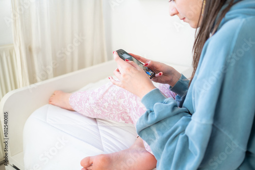 A young woman looks at her mobile phone scrolling on the screen with red painted nails. She is sitting barefoot on a bed, with soft light coming in gently through the window