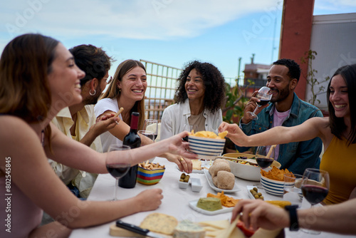 Group of multi-ethnic young friends enjoying meal gathered on rooftop. Happy millennial people having fun drinking red wine and eating celebrating a party event on summer day photo