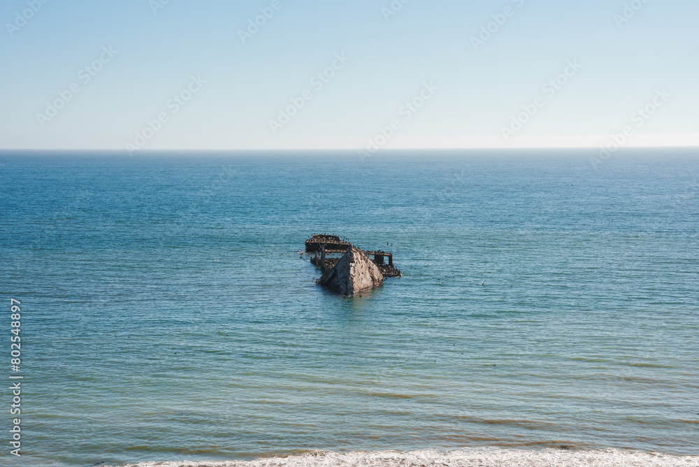 Serene seascape under clear blue skies with calm ocean waters and a shipwreck visible. Rusted and weathered remains suggest age. Tranquil and remote coastline scene. Serene seascape under clear blue skies with calm ocean waters and a shipwreck visible. Rusted and weathered remains suggest age. Tranquil and remote coastline scene.
