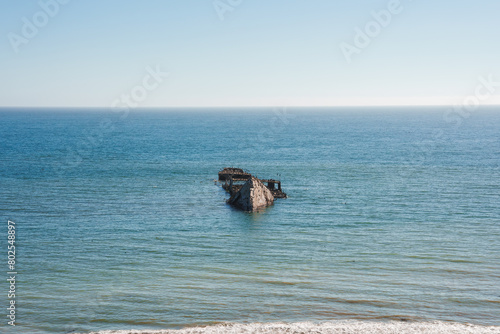 Serene seascape under clear blue skies with calm ocean waters and a shipwreck visible. Rusted and weathered remains suggest age. Tranquil and remote coastline scene.