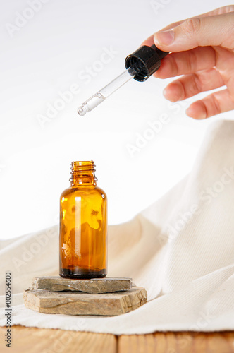 A brown bottle of essential oil sitting on rocks on a natural fiber cloth, on a wooden surface. A hand is holding the dropper to show the transparent liquid inside.
