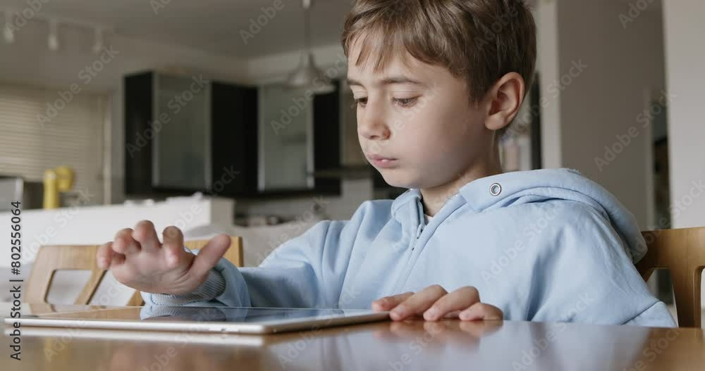 A young boy with blond hair is focused on using a digital tablet while sitting at a wooden table, in a well-lit, spacious living room during the day. A young boy with blond hair is focused on using a digital tablet while sitting at a wooden table, in a well-lit, spacious living room during the day.