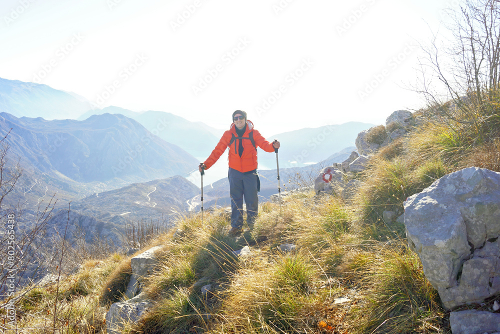 A tourist in a bright jacket with trekking poles poses against the backdrop of a picturesque mountain landscape. Hiking on Mount Orjen in Montenegro