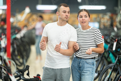 Positive interested young couple walking through specialized bicycle store  looking over variety of modern bikes and gear available  amicably discussing choice for cycling needs
