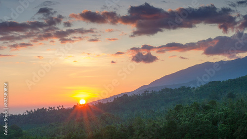 view of hills covered with grass and green trees when the sun has just appeared with a yellowish sky and black clouds on Pergasingan Hill