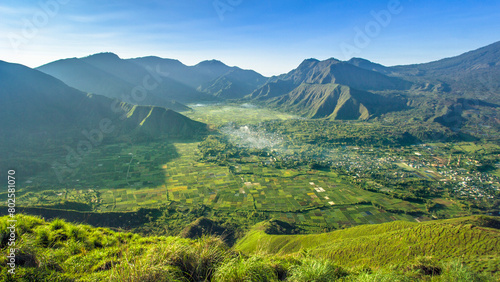 views of several green hills below which there are stretches of rice fields and green farms and residential areas under the hillsides with blue skies on Pergasingan Hill
