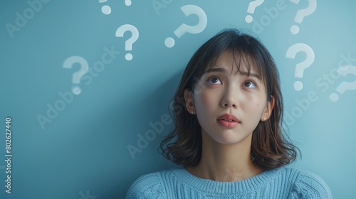 A young woman in a blue sweater is looking up at question marks.