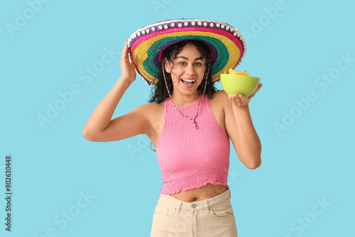 Happy young African-American woman in sombrero hat holding bowl with tasty nachos on blue background