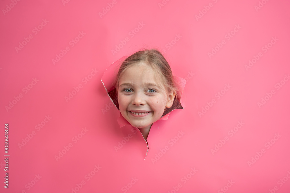 Cute Caucasian girl peeks out of a hole in a paper pink background.