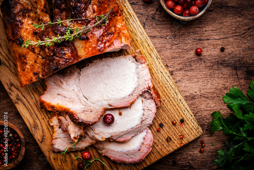 Baked festive pork butt or ham with herbs, spices and cranberries for sauce, served and sliced on cutting board, rustic wooden table background, top view © 5ph