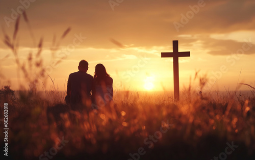 Couple praying together in a field with cross at sunset. 