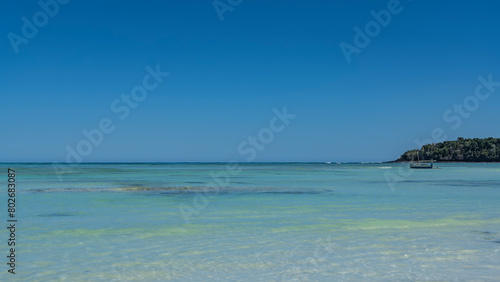 A serene seascape. A wooden boat is visible in the calm aquamarine ocean. The sail is folded. A green island against a clear blue sky. A tropical idyll. Copy space. Madagascar. Nosy Iranja