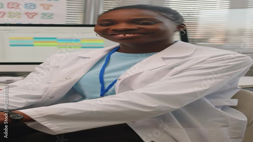 Vertical waist up portrait if young Black female child pediatrician wearing white lab coat sitting at desk in modern doctors office and smiling at camera photo
