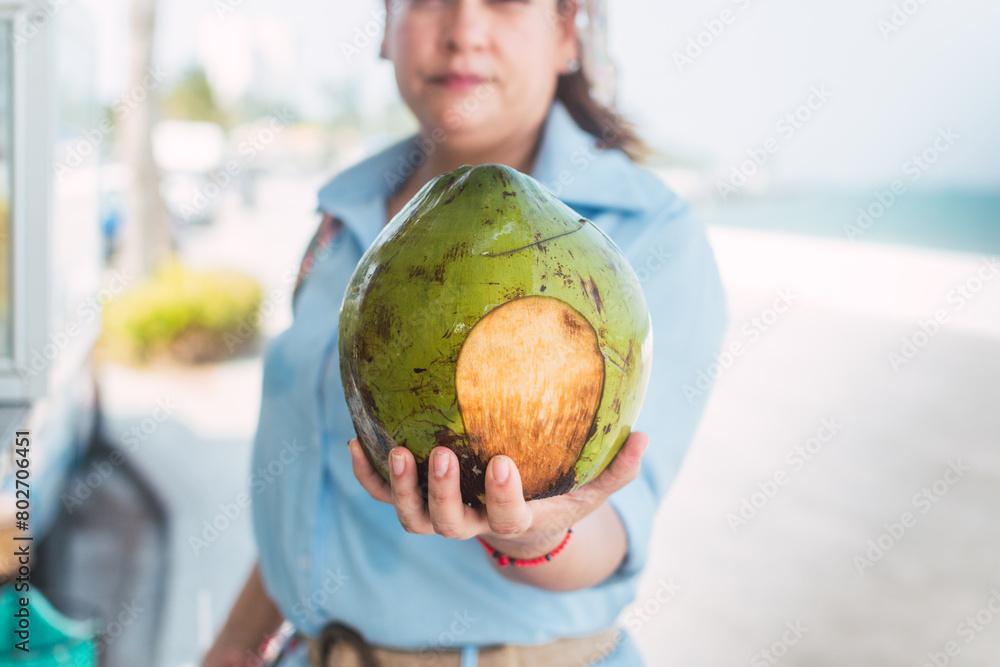 Fototapeta premium Latin woman drinking coconut water on the beach, at a street stall with glasses of tropical fruits.