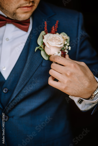 Valmiera, Latvia - August 19, 2023 - Close-up of a groom in a blue suit adjusting a pale rose boutonniere, focusing on the details of his outfit.