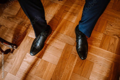 Valmiera, Latvia - August 19, 2023 - Close-up of a groom's polished black leather shoes and dark blue suit pants, standing on a wooden floor.