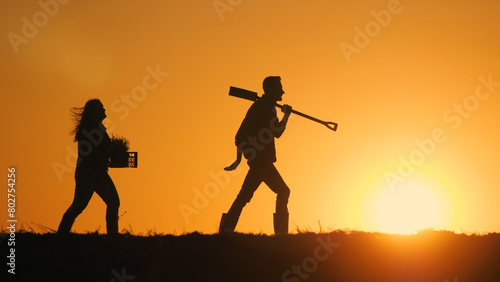 A couple of farmers with equipment walks through a field at sunset.