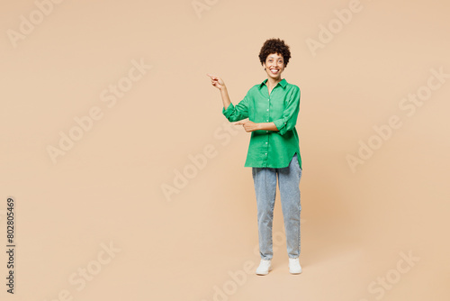 Full body young woman of African American ethnicity wear green shirt casual clothes point index finger aside indicate on area isolated on plain pastel light beige background studio. Lifestyle concept. © ViDi Studio