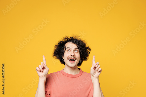 Young smiling happy fun man he wear pink t-shirt casual clothes point index finger overhead on empty blank area workspace isolated on plain yellow orange background studio portrait. Lifestyle concept.