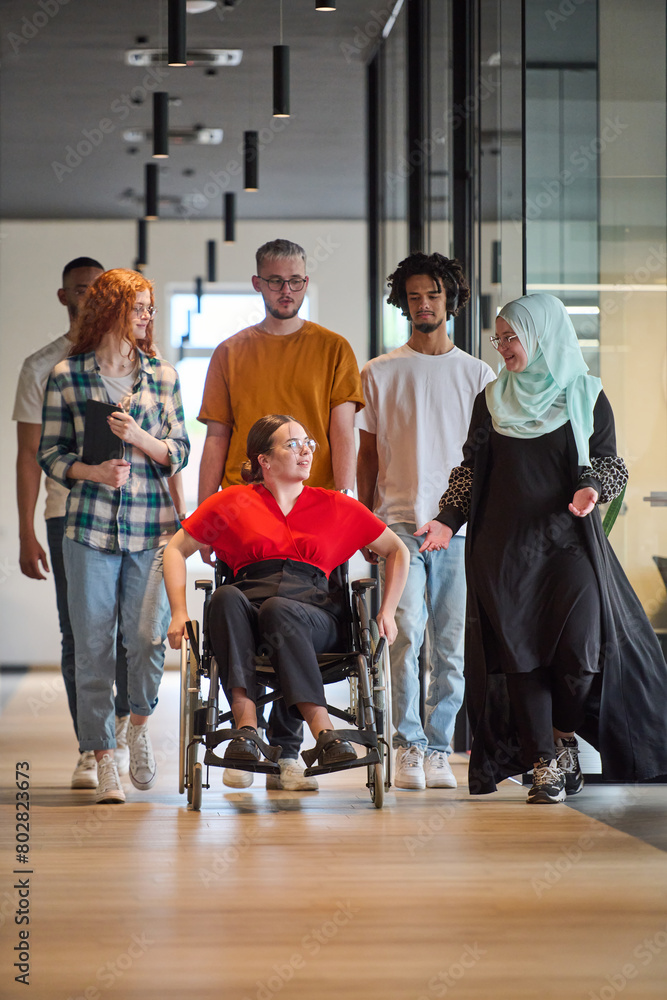 Fototapeta premium A diverse group of young business people walking a corridor in the glass-enclosed office of a modern startup, including a person in a wheelchair and a woman wearing a hijab