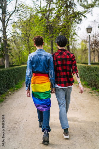 A joyful gay couple with an LGBT flag strolling in a lush park. © Koldo_Studio
