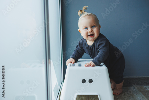 photo of a 7-9 month old child in a home interior. An inquisitive, smiling child explores the world around him. Cute baby stand by the chair. The child learns to stand and walk.