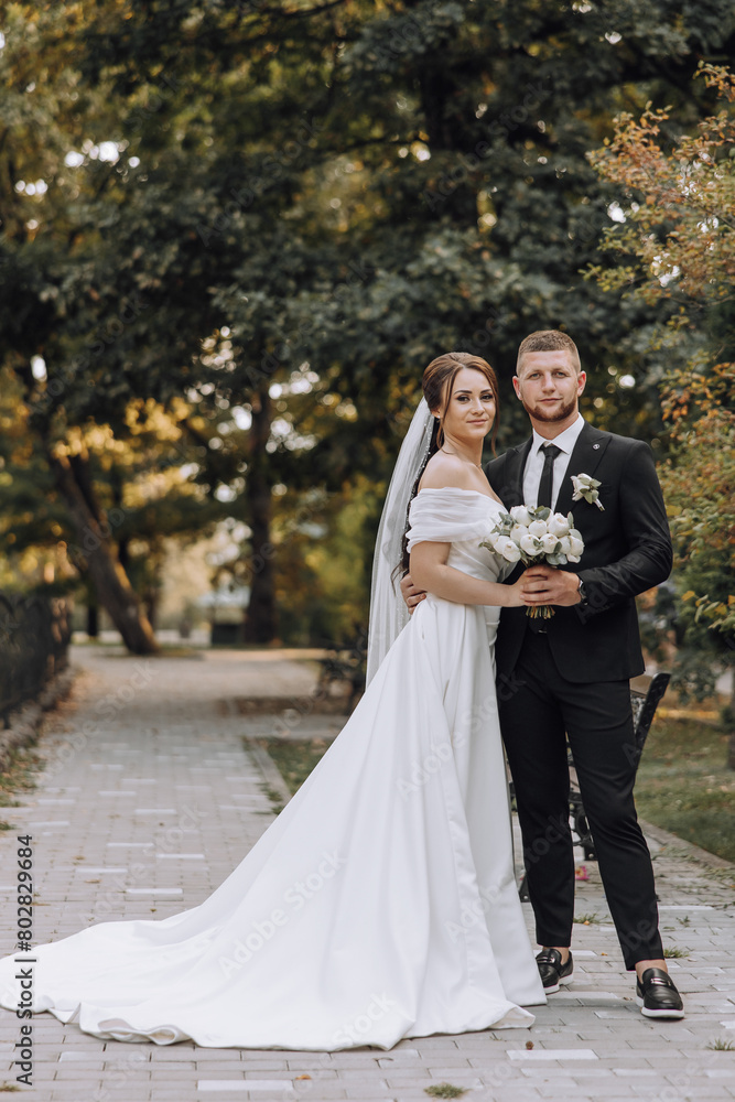 A bride and groom are posing for a picture in a park. The bride is wearing a white dress and the groom is wearing a black suit. They are holding a bouquet and a small bouquet of flowers