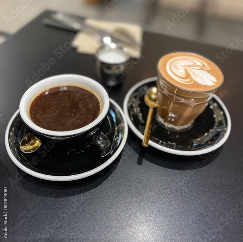 A glass of latte and a cup of coffee on a table with blurry background