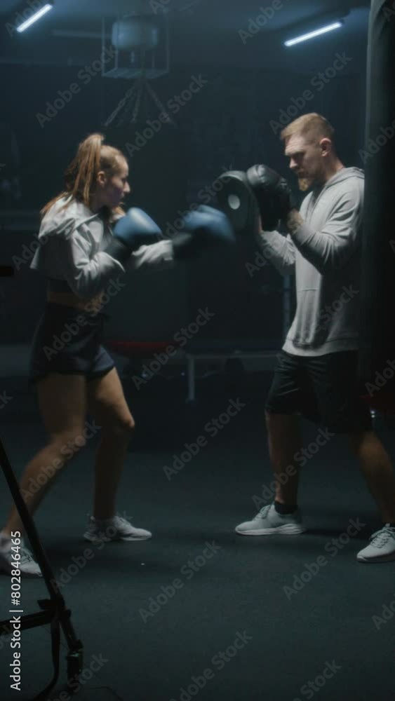 Vertical shot of Female boxer in boxing gloves hits punching mitts and practices fighting techniques with coach in dark boxing gym. Athletic woman exercises and prepares for match. Vertical shot of Female boxer in boxing gloves hits punching mitts and practices fighting techniques with coach in dark boxing gym. Athletic woman exercises and prepares for match.