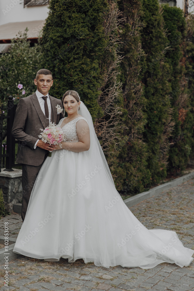 A bride and groom are posing for a picture in front of a bush. The bride is wearing a white dress and the groom is wearing a suit. The couple is holding a bouquet and a bouquet of flowers