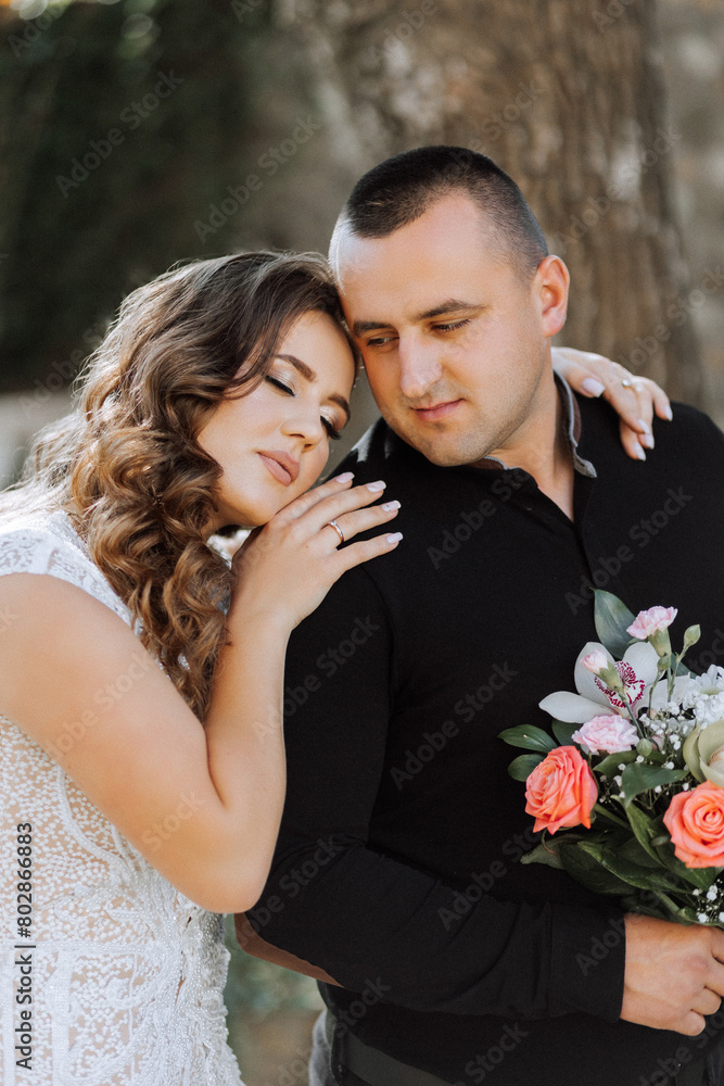 A man and a woman are hugging each other. The woman is wearing a wedding dress and the man is wearing a black shirt. They are holding a bouquet of flowers A man and a woman are hugging each other. The woman is wearing a wedding dress and the man is wearing a black shirt. They are holding a bouquet of flowers