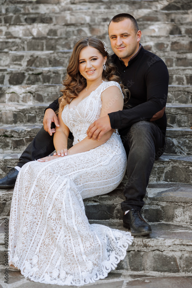 A man and woman are sitting on a set of stairs, smiling for the camera. The woman is wearing a white dress and the man is wearing a black shirt and jeans. Scene is happy and romantic
