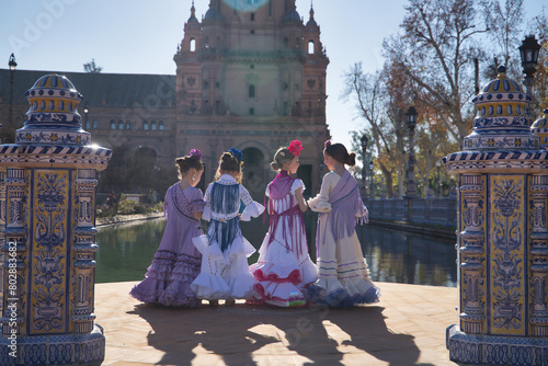 Four girls dancing flamenco, with their backs turned and talking to each other, in typical flamenco costumes next to a lake in a beautiful square in Seville. Concept dance, flamenco, typical Spanish.