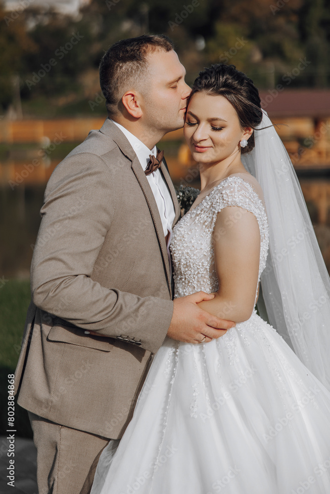 Naklejka premium A bride and groom are kissing each other's cheeks. The bride is wearing a white dress and the groom is wearing a suit