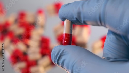 Red and white capsule in human hand in medical glove close-up on with a blurred background of a bunch of medicines pills. Healthcare and pharmacy industry concept. photo