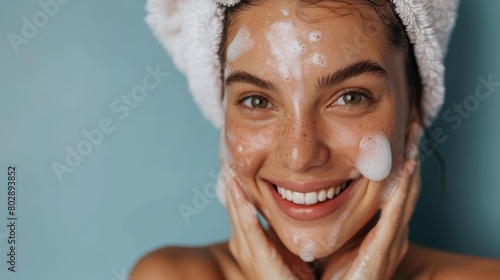 Smiling woman with face wash foam and towel on her head. Close-up studio portrait on blue background. photo