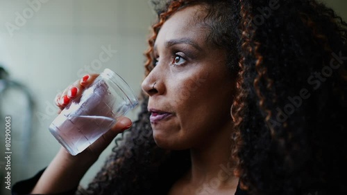 One thoughtful black middle-aged woman drinking water while gazing in the distance with pensive emotion. Contemplative latina person of African descent hydrating herself photo