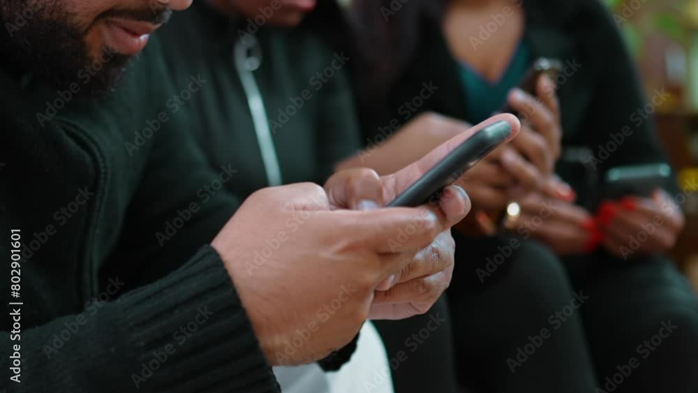 One young black latin man typing message on cellphone device with blurred friends on background. 20s person seated on couch using modern technology, holding phone adn staring at screen One young black latin man typing message on cellphone device with blurred friends on background. 20s person seated on couch using modern technology, holding phone adn staring at screen